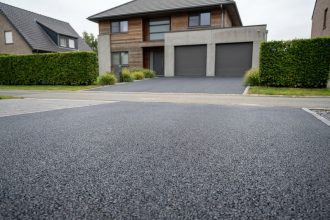 Allée de maison en enrobé bitumineux gris anthracite menant à un garage double, entourée de haies vertes soignées
